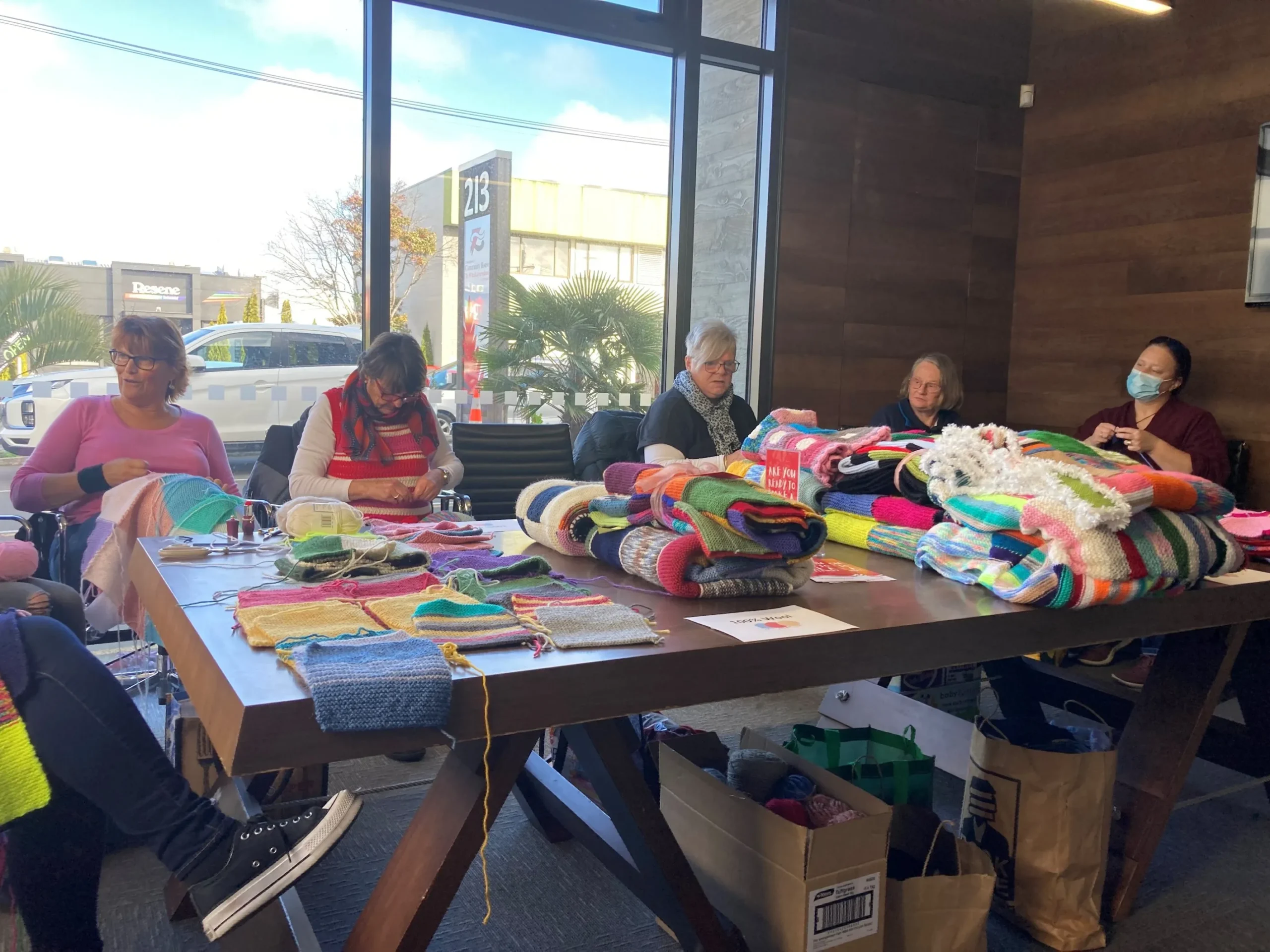 Knitters sitting around a table piled high with knitted blankets and peggy squares