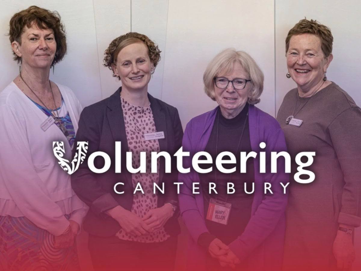Women standing and smiling at the camera, representing the team from Volunteering Canterbury. The Volunteering Canterbury logo is displayed prominently across the image with a red-to-purple gradient overlay.